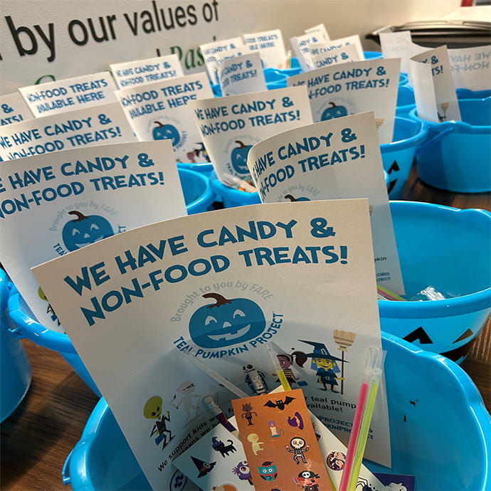 Blue plastic buckets filled with flyers that read “We have candy & non‑food treats!” along with small toys and stickers, representing the Teal Pumpkin Project.