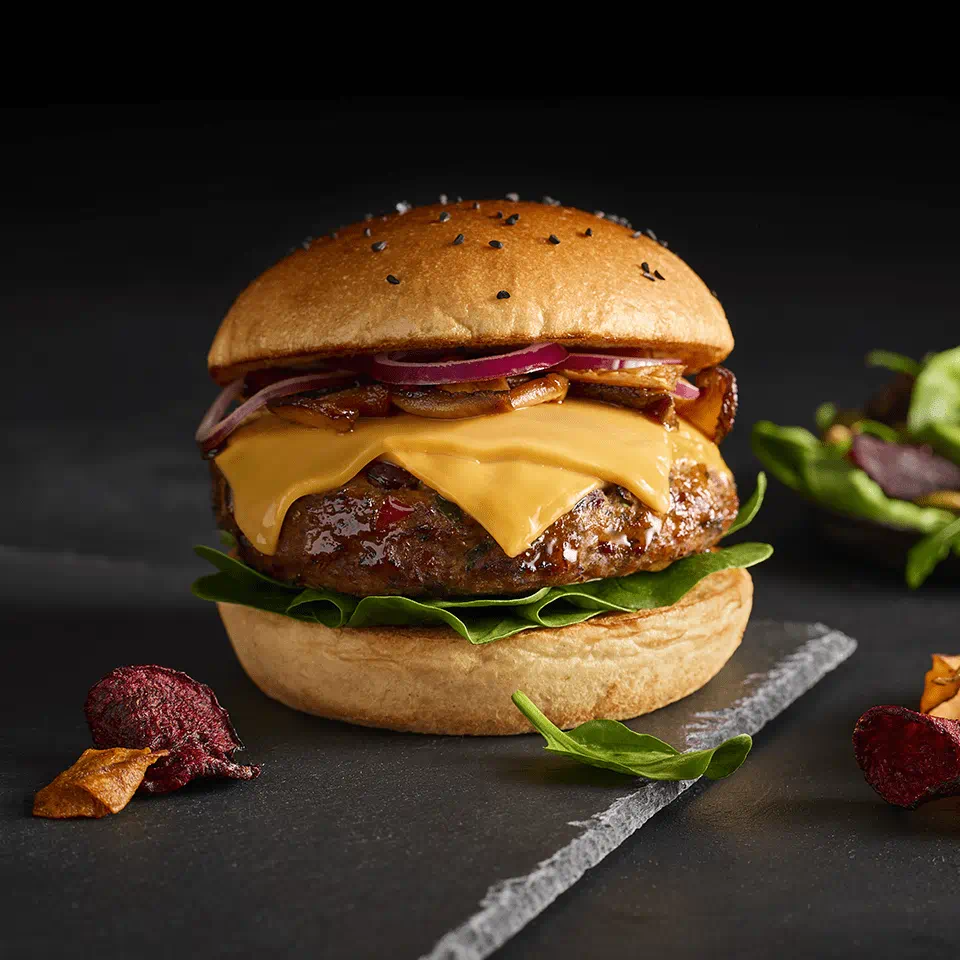 A plant-based burger with melted dairy free cheese slices, red onions, and greens in a sesame-topped bun, placed on a slate board with vegetable chips.