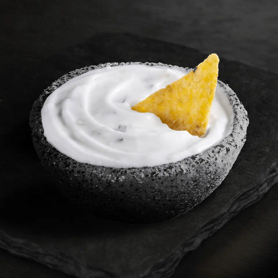 A stone-textured bowl of dairy free dip with a tortilla chip dipped into it, placed on a dark slate surface.