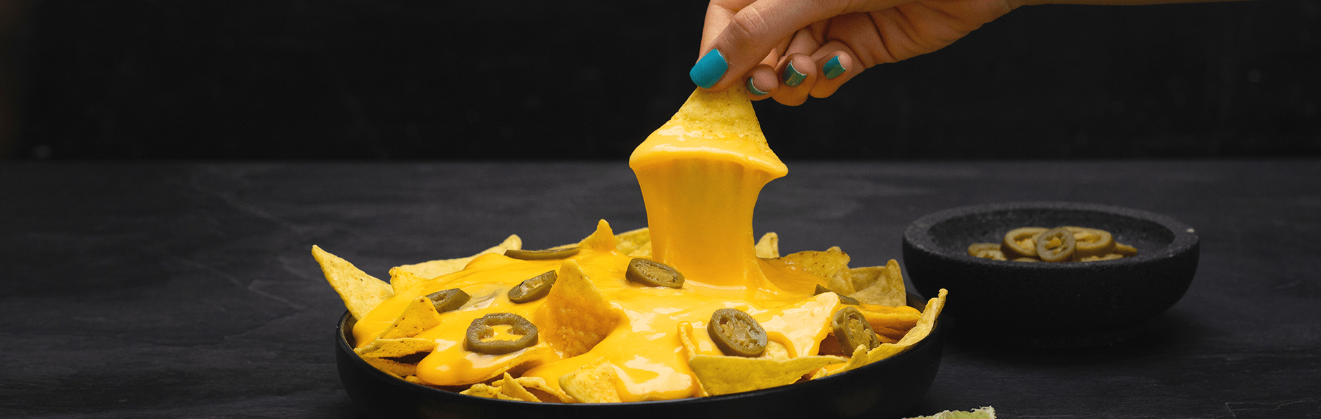 A hand dips a tortilla chip into warm, melted Violife dairy free cheese sauce, with nachos and jalapeño slices visible underneath.