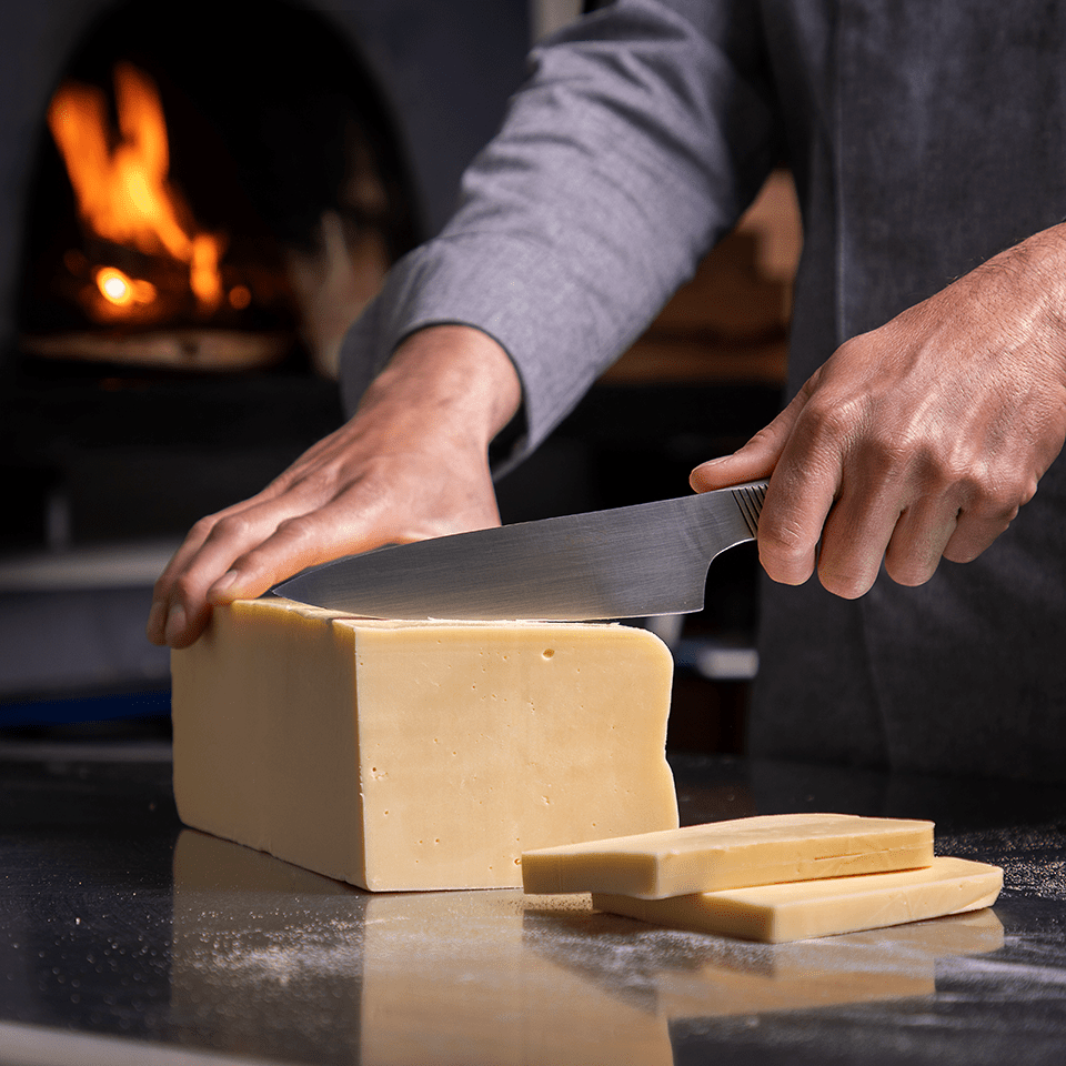 Hands slicing a large block of dairy-free cheese on a countertop with a chef’s knife. Two rectangular slices are placed in front and a wood-fired oven glows in the background.