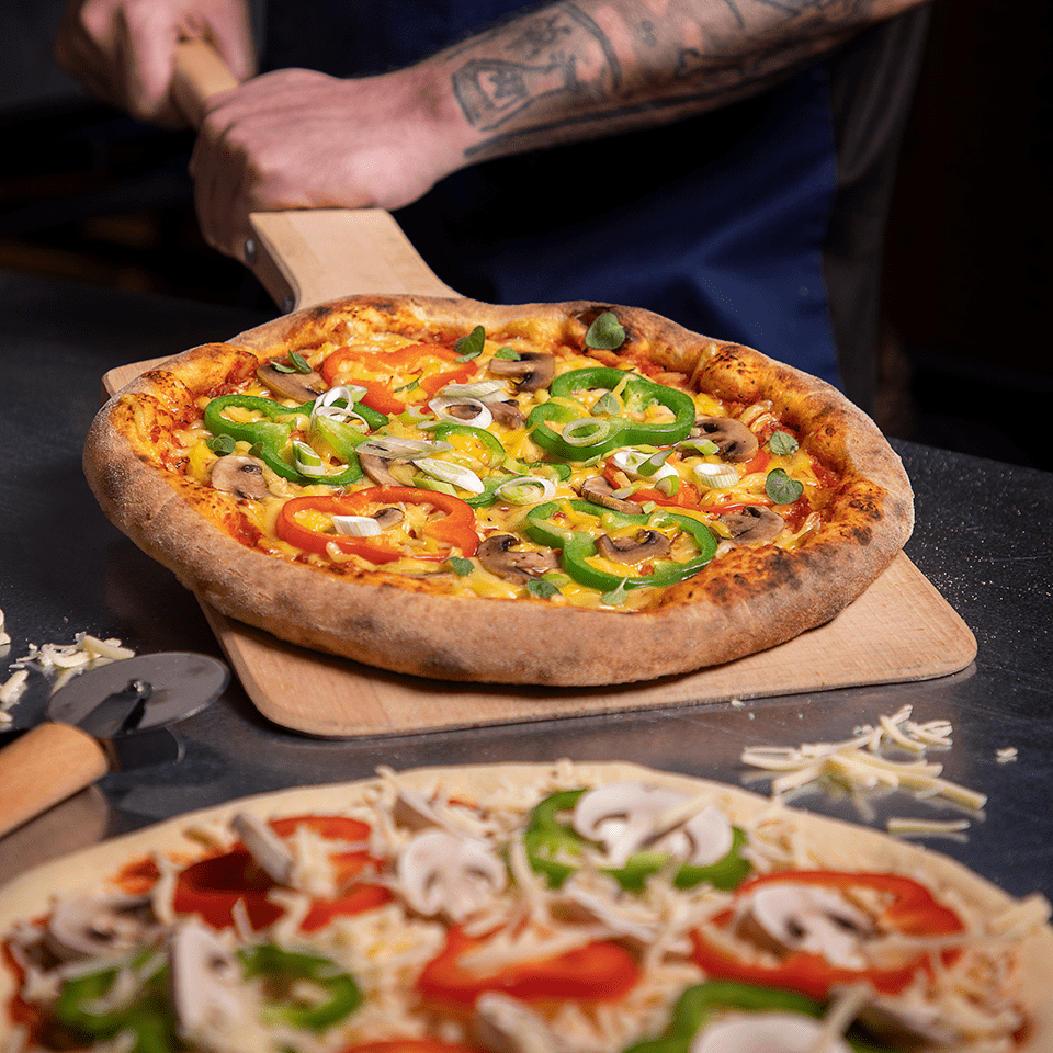 A freshly baked pizza topped with green and red bell peppers, mushrooms, and melted Violife. Another uncooked pizza with same toppings is visible in the foreground on a counter, along with a pizza cutter and scattered dairy free cheese shreds.
